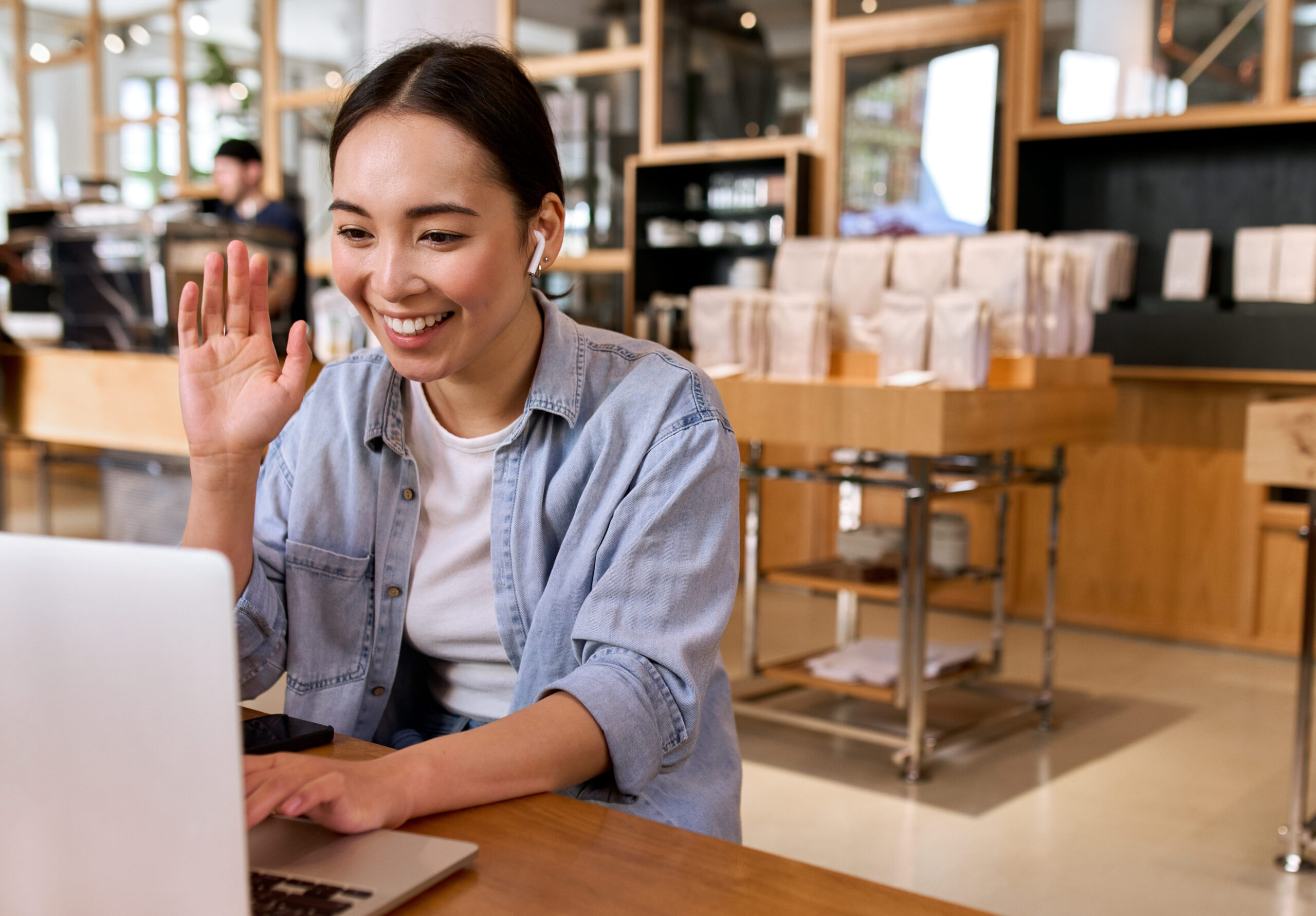 Person seated at a café table smiling and waving during a video call on a laptop, with earbuds in.